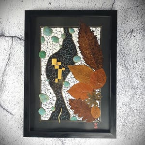 May include: A framed artwork featuring a mosaic of small, white tiles with a black, abstract design. The mosaic is accented with dried leaves in shades of brown, orange, and green. The artwork is signed in red with the character "草".
