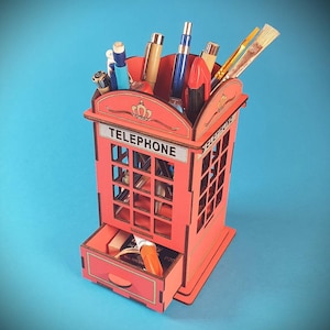 May include: A red wooden desk organizer shaped like a British telephone booth. The organizer has a drawer and is filled with pens, pencils, and paintbrushes. The word "TELEPHONE" is displayed on the front.