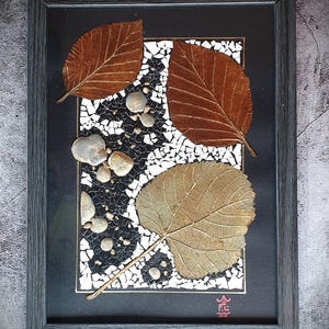 Può includere: Un'opera d'arte in mosaico incorniciata con foglie secche in tonalità di marrone e oro, su uno sfondo nero con accenti bianchi e argentati. L'opera è incorniciata in una cornice grigio scuro.