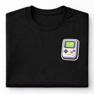 May include: Black t-shirt featuring a colorful, cartoon-style graphic of a handheld video game console. The console has a green screen, purple buttons, and a white border. A classic design for gamers.