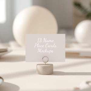 May include: A light beige place card held by a small, round, concrete-like holder. The card has the text "13 Name Place Cards Mockups" in a soft, elegant font. The background features soft, blurred shapes and shadows.