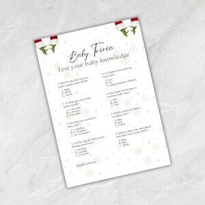 May include: A white baby trivia game sheet with red and green accents. The text reads "Baby Trivia" and "Test your baby knowledge." Questions cover baby development and facts. Decorative snowflakes and Christmas-themed accents.