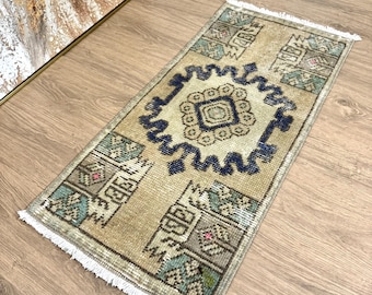 May include: A small, rectangular rug with a beige background and a blue and green geometric pattern. The rug has a fringe border.