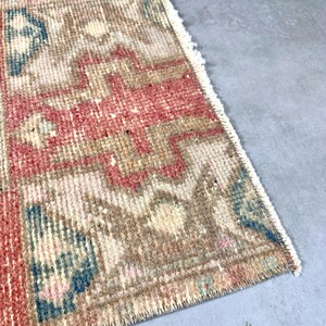 May include: A small, rectangular rug with a faded, vintage appearance. The rug features a geometric pattern in shades of red, beige, and blue. The edges are fringed, adding to its aged aesthetic. The rug is likely made of woven material.