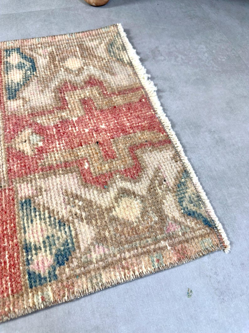 May include: A small, rectangular rug with a faded, vintage appearance. The rug features a geometric pattern in shades of red, beige, and blue. The edges are fringed, adding to its aged aesthetic. The rug is likely made of woven material.