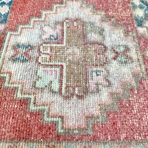 May include: Close-up of a small, rectangular rug with a geometric pattern. The rug features a central design in shades of brown, pink, and beige, surrounded by a red border. The rug's design includes a series of stepped shapes and cross-like motifs.
