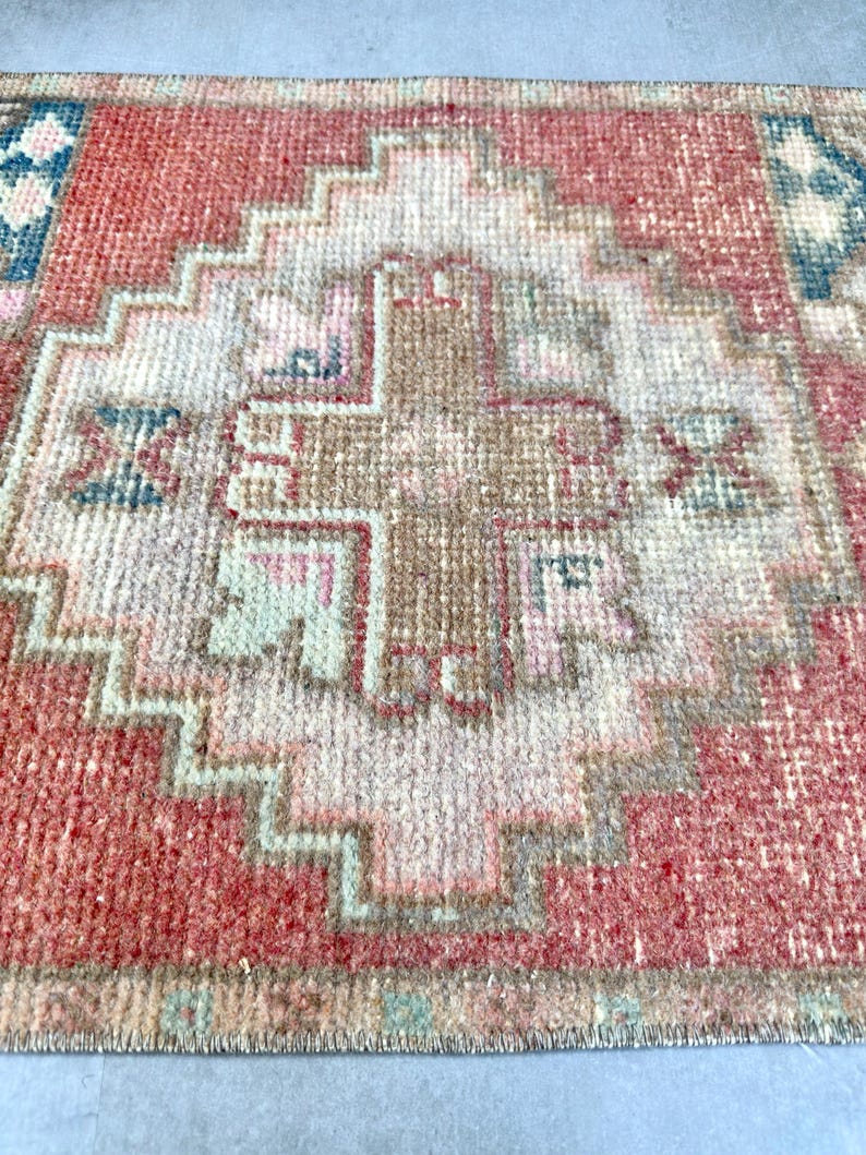 May include: Close-up of a small, rectangular rug with a geometric pattern. The rug features a central design in shades of brown, pink, and beige, surrounded by a red border. The rug's design includes a series of stepped shapes and cross-like motifs.
