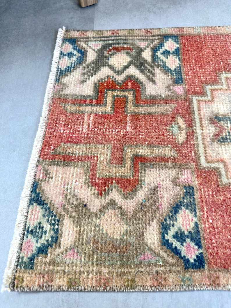 May include: A small, rectangular rug with a geometric pattern. The rug features a combination of red, beige, blue, and gray colors. The design includes repeating shapes and patterns, with a fringed edge along one side. The rug appears to be made of woven material.