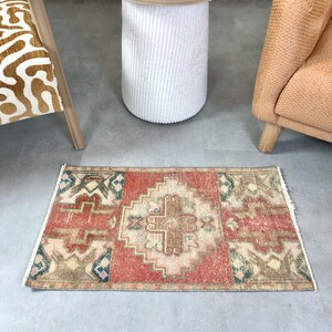 May include: A rectangular, hand-knotted rug with a central geometric design in shades of red, beige, and brown. The rug features a border with blue and green accents. The rug is placed on a gray floor.