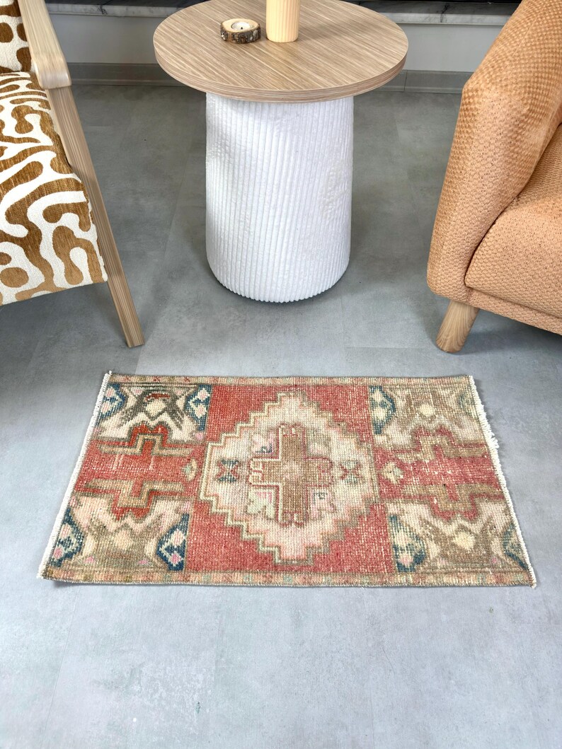 May include: A rectangular, hand-knotted rug with a central geometric design in shades of red, beige, and brown. The rug features a border with blue and green accents. The rug is placed on a gray floor.
