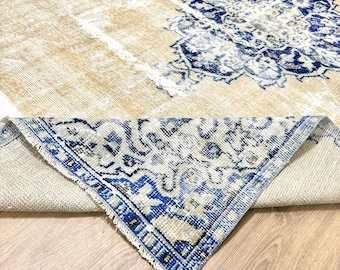 May include: A distressed beige rug featuring a large blue and white medallion design. The rug's corner is folded up, revealing a detailed border with blue and white patterns. The rug is placed on a light wood surface.