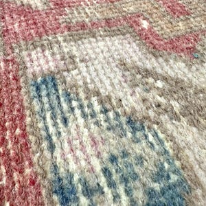 May include: Close-up of a woven rug with a textured surface. The rug features a pattern of red, blue, and beige hues. The design appears to be a traditional style, with visible knots and variations in the fibers.
