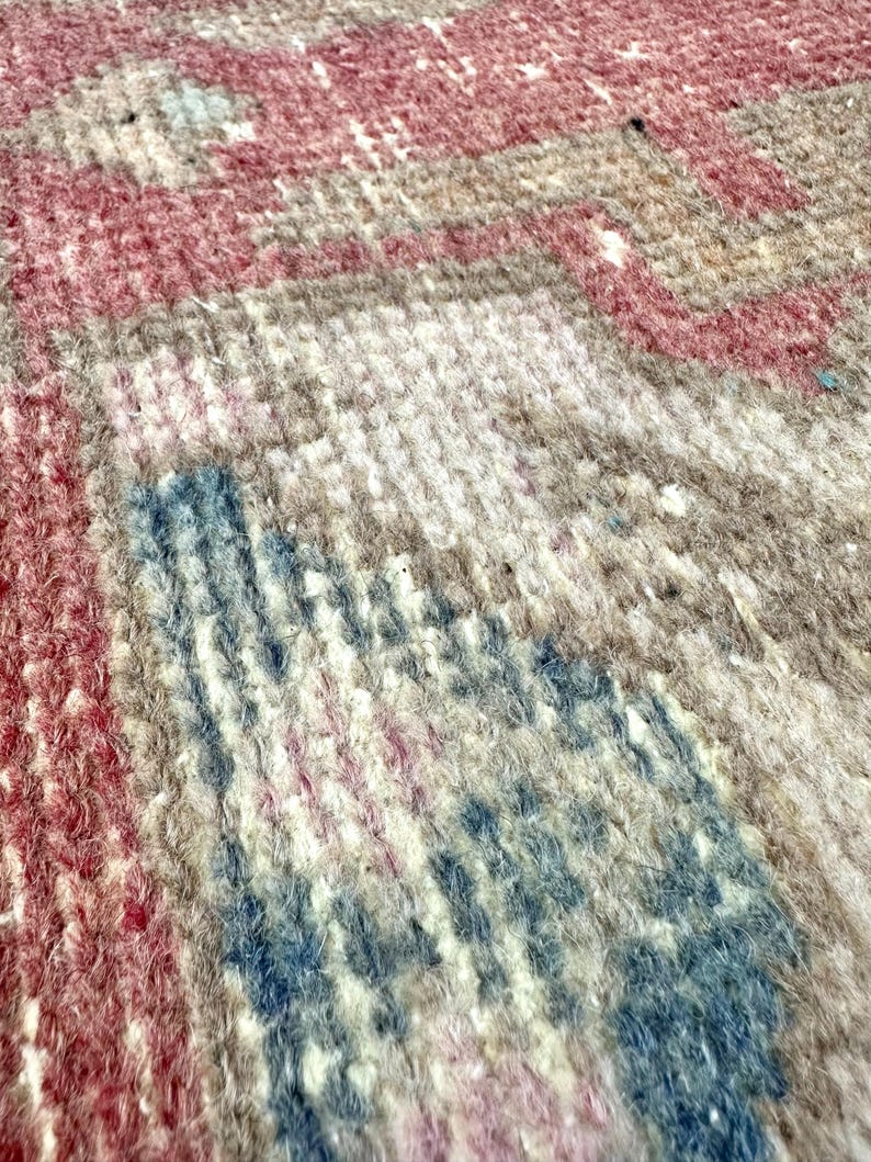 May include: Close-up of a woven rug with a textured surface. The rug features a pattern of red, blue, and beige hues. The design appears to be a traditional style, with visible knots and variations in the fibers.