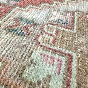 May include: Close-up of a hand-woven rug with a geometric pattern. The rug features a mix of colors, including red, pink, beige, and blue. The texture is visible, showing the individual threads and knots.