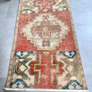 May include: A rectangular, hand-woven rug with a central red field and intricate geometric patterns in beige, blue, and brown. The rug has a fringed edge and a vintage, distressed appearance, suitable for a hallway or entryway.