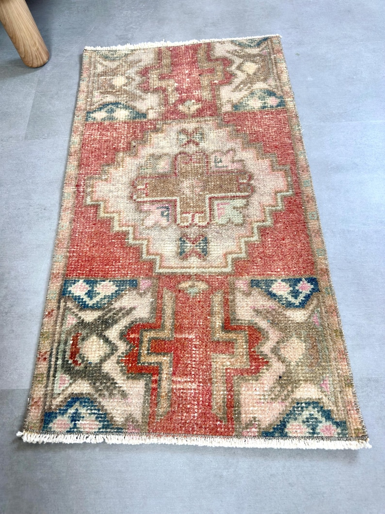 May include: A rectangular, hand-woven rug with a central red field and intricate geometric patterns in beige, blue, and brown. The rug has a fringed edge and a vintage, distressed appearance, suitable for a hallway or entryway.