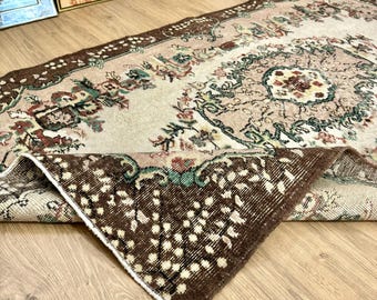 May include: A vintage rug with a cream background and a brown border. The rug has a floral pattern with green, brown, and red accents. The rug is folded in half, showing the underside.