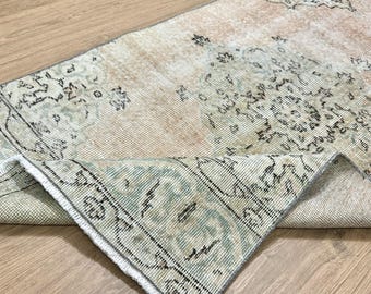 May include: A vintage rug with a faded, light brown background and a green and black floral pattern. The rug is folded in half, showing the underside.