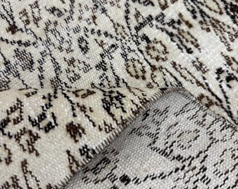 May include: Close-up of a white rug with a brown and black geometric pattern. The rug is folded in half, showing the underside of the fabric.