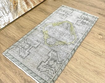 May include: A rectangular rug with a distressed appearance, featuring a faded floral design in grey, green, and white. The rug has a textured surface and a fringed edge. It is positioned on a wooden floor.