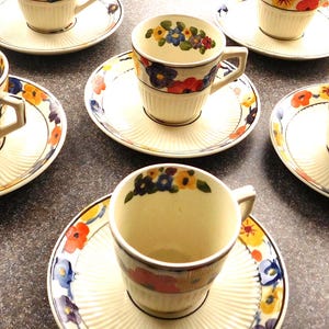 May include: Set of six cream-colored teacups and saucers. Each cup and saucer features a floral design with red, yellow, and blue flowers. The saucers have a ridged pattern, and the cups have a simple handle.