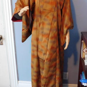 May include: A full-length kimono featuring a textured pattern in shades of burnt orange, mustard yellow, and muted brown. The garment has wide, flowing sleeves, a structured collar, and a deep red lining visible at the interior of the sleeve cuffs.
