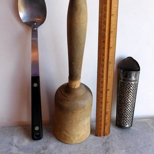 May include: A wooden kitchen mallet with a rounded head, a metal grater, and a silver spoon with a black handle. The items are arranged on a white surface next to a wooden ruler.