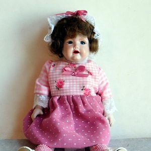 May include: A vintage porcelain doll with brown curly hair, blue eyes, and a pink gingham dress with rose accents. The doll wears a white lace bonnet and matching pink socks and shoes. The doll is sitting down.