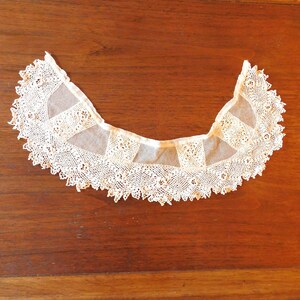 May include: A white lace collar with a scalloped edge. The collar is made of delicate, intricate lace and has a vintage look.