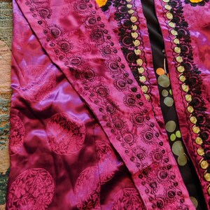 K&ouml;nnte beinhalten: Ein lebendiger magentafarbener Robe mit komplizierten schwarzen, gestickten Blumenmustern an den R&auml;ndern. Die Robe hat einen satinartigen Glanz und einen kontrastierenden schwarzen Stoff mit bunten Tupfen. Das Design ist reich und detailliert.