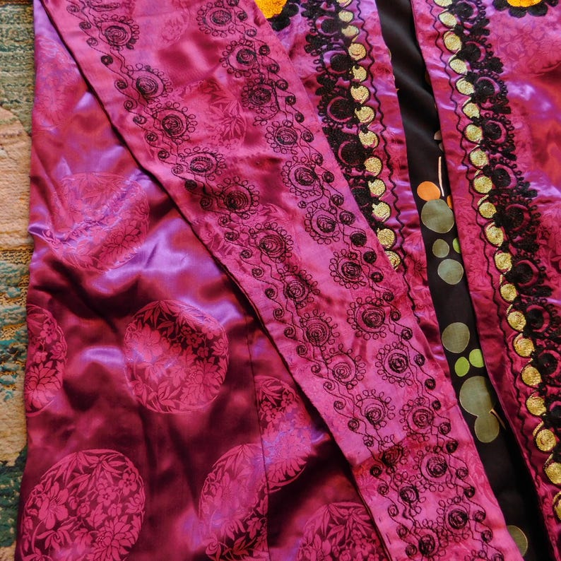 K&ouml;nnte beinhalten: Ein lebendiger magentafarbener Robe mit komplizierten schwarzen, gestickten Blumenmustern an den R&auml;ndern. Die Robe hat einen satinartigen Glanz und einen kontrastierenden schwarzen Stoff mit bunten Tupfen. Das Design ist reich und detailliert.