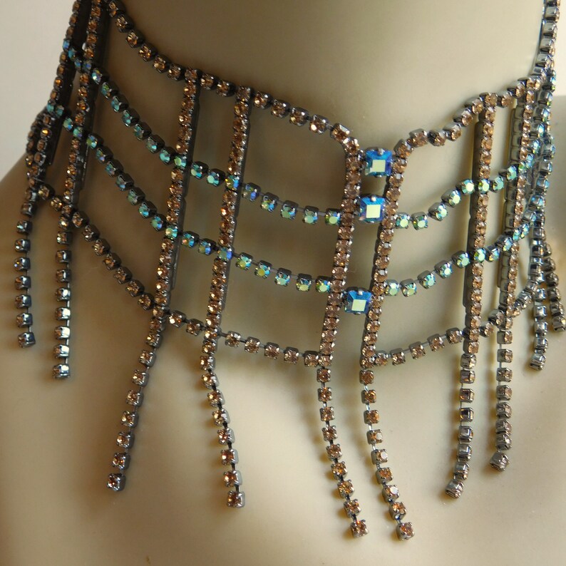 May include: A silver choker necklace with a geometric design made of small, clear and blue rhinestones. The necklace has multiple strands of rhinestones that hang down from the choker.