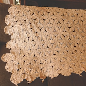 May include: A crocheted throw blanket with a geometric pattern in a cream color. The blanket has a scalloped edge and fringe.