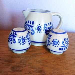 May include: A ceramic pitcher and two salt shakers. The set is white with blue floral designs and trim. The pitcher has a handle and spout. The salt shakers are egg-shaped with holes on top. The set is on a wooden surface.