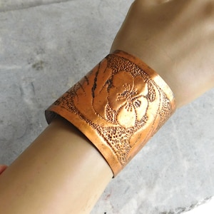 Puede incluir: Pulsera ancha de cobre con un diseño floral en relieve. La pulsera es de color marrón rojizo cálido y presenta un patrón detallado de flores y hojas. La pulsera mide aproximadamente 5 cm de ancho.