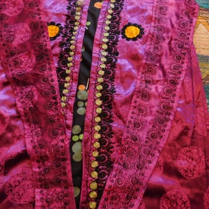 K&ouml;nnte beinhalten: Ein magentafarbenes Kleidungsst&uuml;ck mit aufwendiger schwarzer und goldener Stickerei. Das Kleidungsst&uuml;ck hat ein Blumenmuster und einen kontrastierenden schwarzen Einsatz mit gr&uuml;nen und orangenen Kreisen. Gelbe Blumenakzente sind ebenfalls sichtbar.