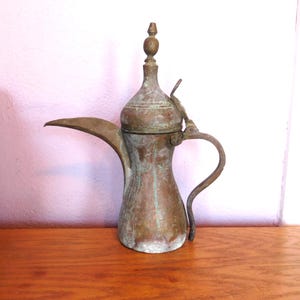 May include: Antique copper-toned coffee pot with a long spout, curved handle, and a decorative finial on the lid. The pot shows signs of age with a patina and some green oxidation. It is likely used for serving coffee or tea.
