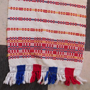 May include: A white woven table runner with red, blue, and yellow stripes. The runner has a fringe trim in red, white, and blue.