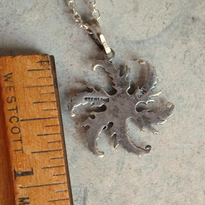 Op de afbeelding: Een zilveren ketting met een hanger met een gedetailleerd, ingewikkeld ontwerp dat lijkt op een gestileerde bloem of stervorm. De hanger is aan de achterkant gemarkeerd met "STERLING". De ketting is ongeveer 45 cm lang.