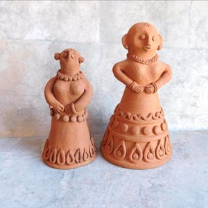 May include: Two terracotta clay figurines of stylized female figures. The figures have rounded bodies, detailed necklaces, and patterned skirts. The figures are standing, with their arms crossed over their chests. The figurines are a warm, earthy tone.