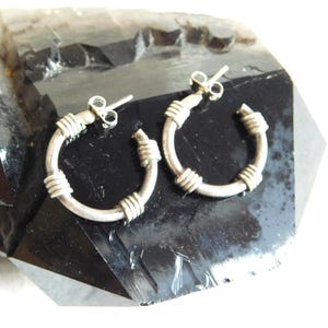 May include: A pair of silver hoop earrings with a wrapped wire design. The earrings are circular, with a post for piercing. The wire is wrapped around the hoop in several places, creating a textured look. The earrings are displayed on a dark, shiny surface.