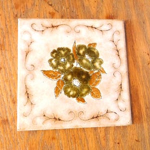 May include: A square ceramic tile featuring a central floral motif with dark olive green petals and orange leaves. The background is a pale cream color with a delicate, thin brown vine-like border pattern framing the edges of the tile surface.