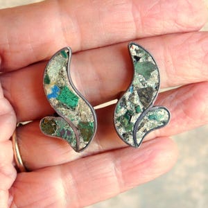 以下が含まれることがあります： シルバーカラーのメタルフレームの抽象的なイヤリングのペア。イヤリングは、緑、青、ベージュの石の破片を使ったモザイクデザインが特徴です。イヤリングは、曲線的で有機的な形状をしています。