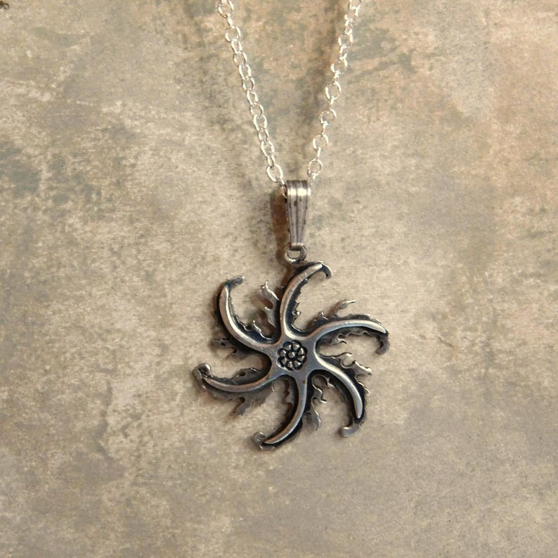Op de afbeelding: Een zilveren ketting met een hanger in de vorm van een gestileerde ster met een gedetailleerd midden. De hanger is bevestigd aan een zilveren ketting.