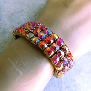 Op de afbeelding: Een brede armband met drie rijen ronde, iriserende kristallen in roze, oranje en gele tinten, gezet in een goudkleurige metalen zetting, wat zorgt voor een levendige, veelzijdige uitstraling rondom de pols van de drager.
