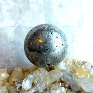 Pode incluir: Uma esfera de pirite, também conhecida como ouro dos tolos, com uma cor cinza prateada metálica e uma superfície texturizada. A esfera repousa sobre uma base de cristais de quartzo claros e amarelos, criando um contraste.