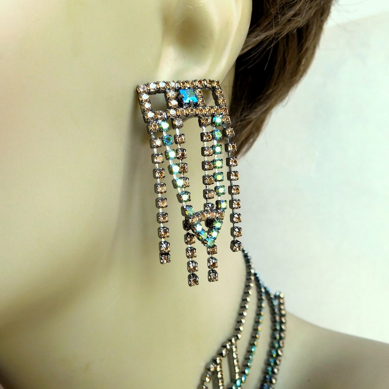 May include: A pair of long, dangling earrings with a geometric design. The earrings are made of metal and feature a cascade of iridescent crystals in shades of blue and brown.