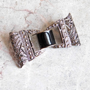 May include: A silver-toned bow-shaped brooch with a black center piece. The brooch is decorated with a geometric pattern of small stones.