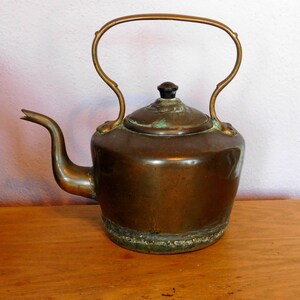 May include: A vintage copper kettle with a tarnished finish and a black knob on the lid. The kettle has a curved handle and a spout.