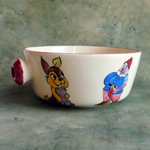 Può includere: Una ciotola in ceramica bianca con un bordo rosso e un manico. La ciotola è decorata con illustrazioni dipinte a mano di un coniglio e un nano. Il coniglio è marrone con la pelliccia gialla e il nano indossa un abito blu e rosso.